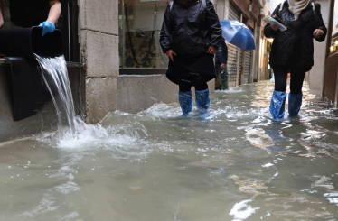 worker empties the water from her shop with a bucket and other people with waterproof gaiters as they walk in the street completely flooded